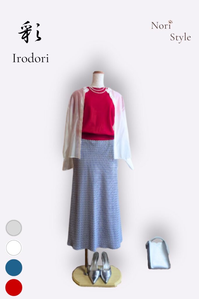 レッドのニットにブルー柄のマーメードスカートを合わせ、ホワイトのシアーブラウスを肩に掛けた春夏コーデ。足元とバッグはシルバーで統一し、鮮やかさと軽やかさが共存する大人のスタイル。