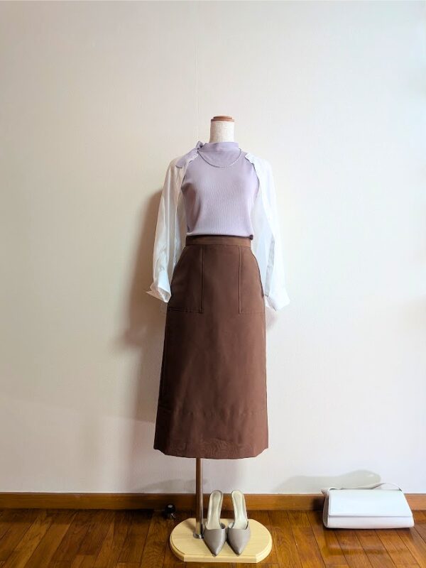 白の背景に佇む、涼やかで優しい大人のフェミニンコーディネート。こっくりとしたチョコレートブラウンのタイトスカートに、甘さ控えめな知的なラベンダーのカットソーを合わせています。上からホワイトシアーブラウスを軽やかに羽織らせ、手元にはオフホワイトのバッグ、足元にはなじみの良いトープのパンプスを優しく添えています。